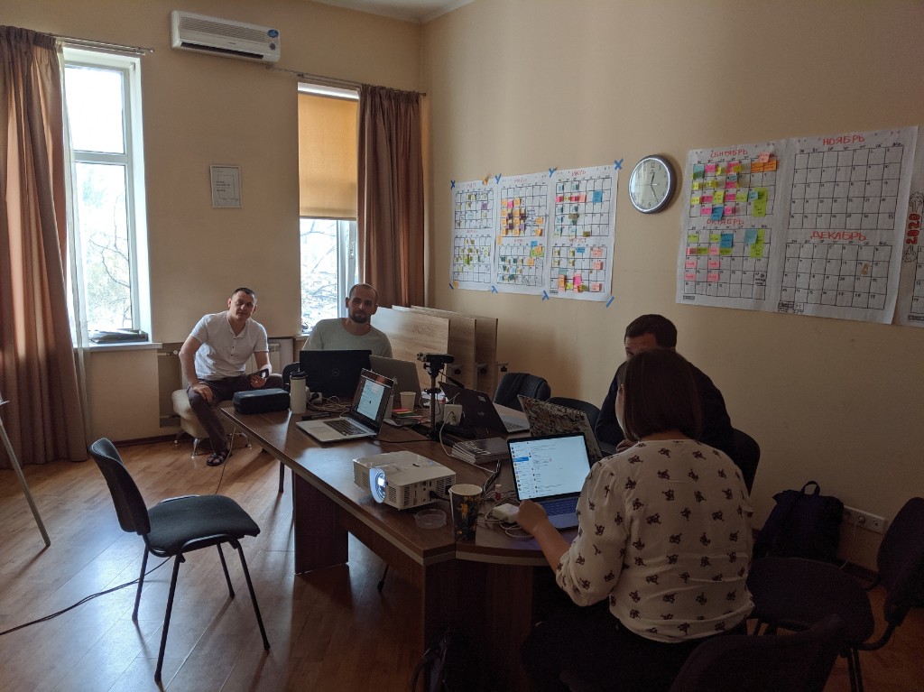 A small team working on laptops around a table during an on-site training session, with planning calendars on the wall.